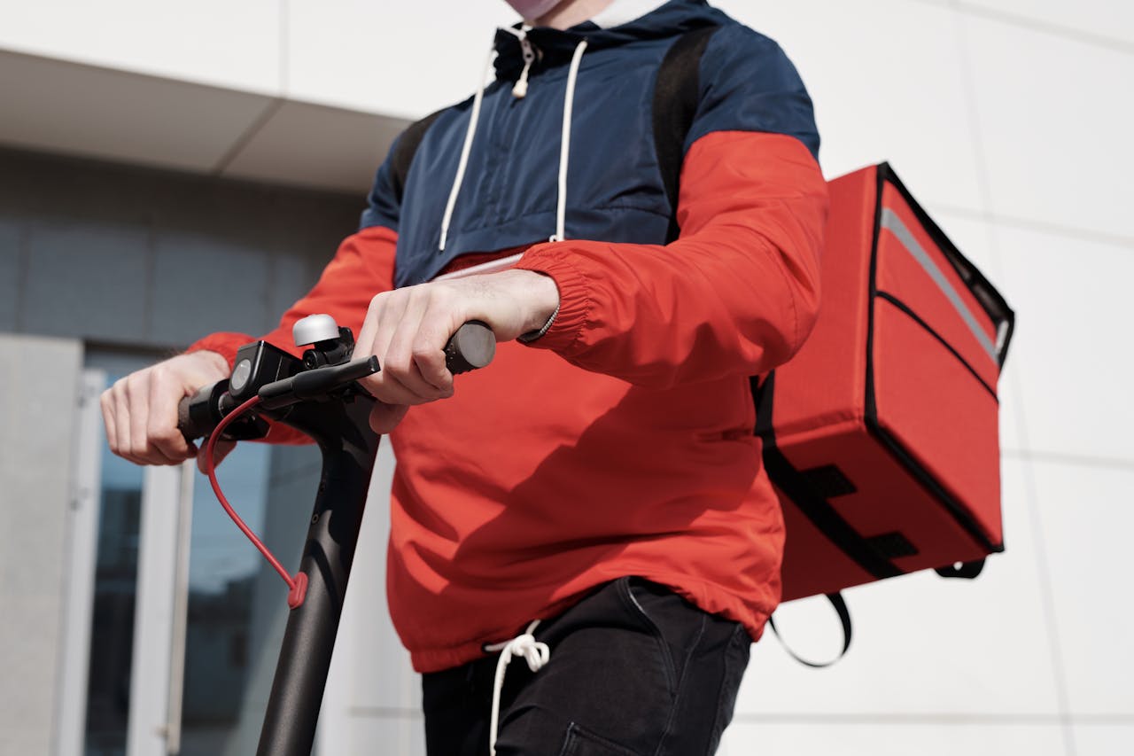 A food delivery person rides a scooter outdoors, wearing a red jacket and carrying a delivery bag.