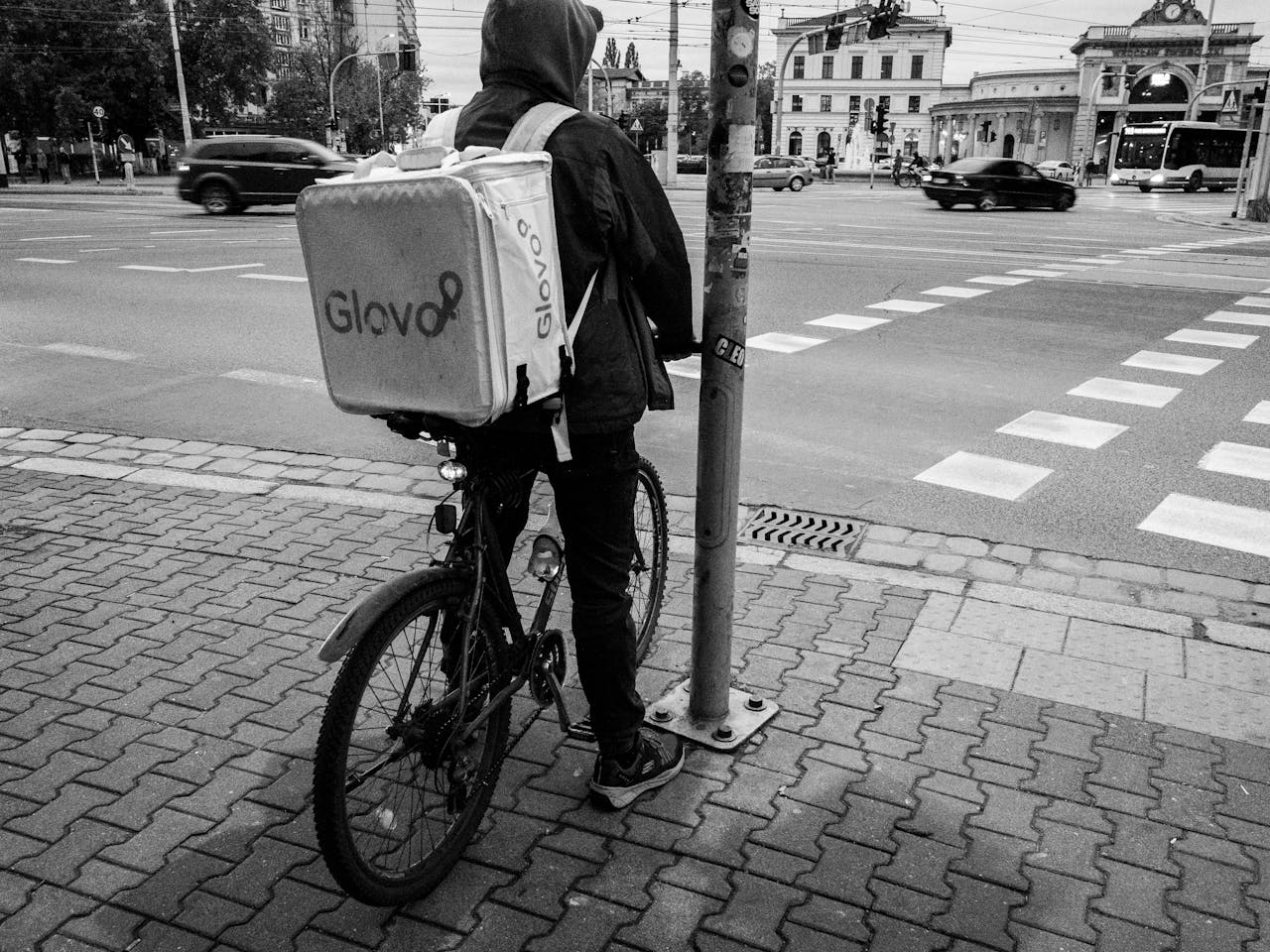 A delivery cyclist with a Glovo bag waits by a city street. Urban logistics in action.