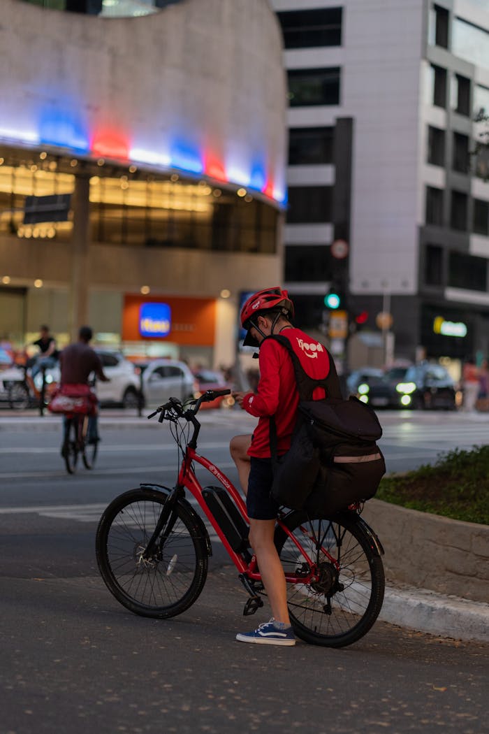 Food delivery cyclist pauses in city street at dusk with illuminated buildings.